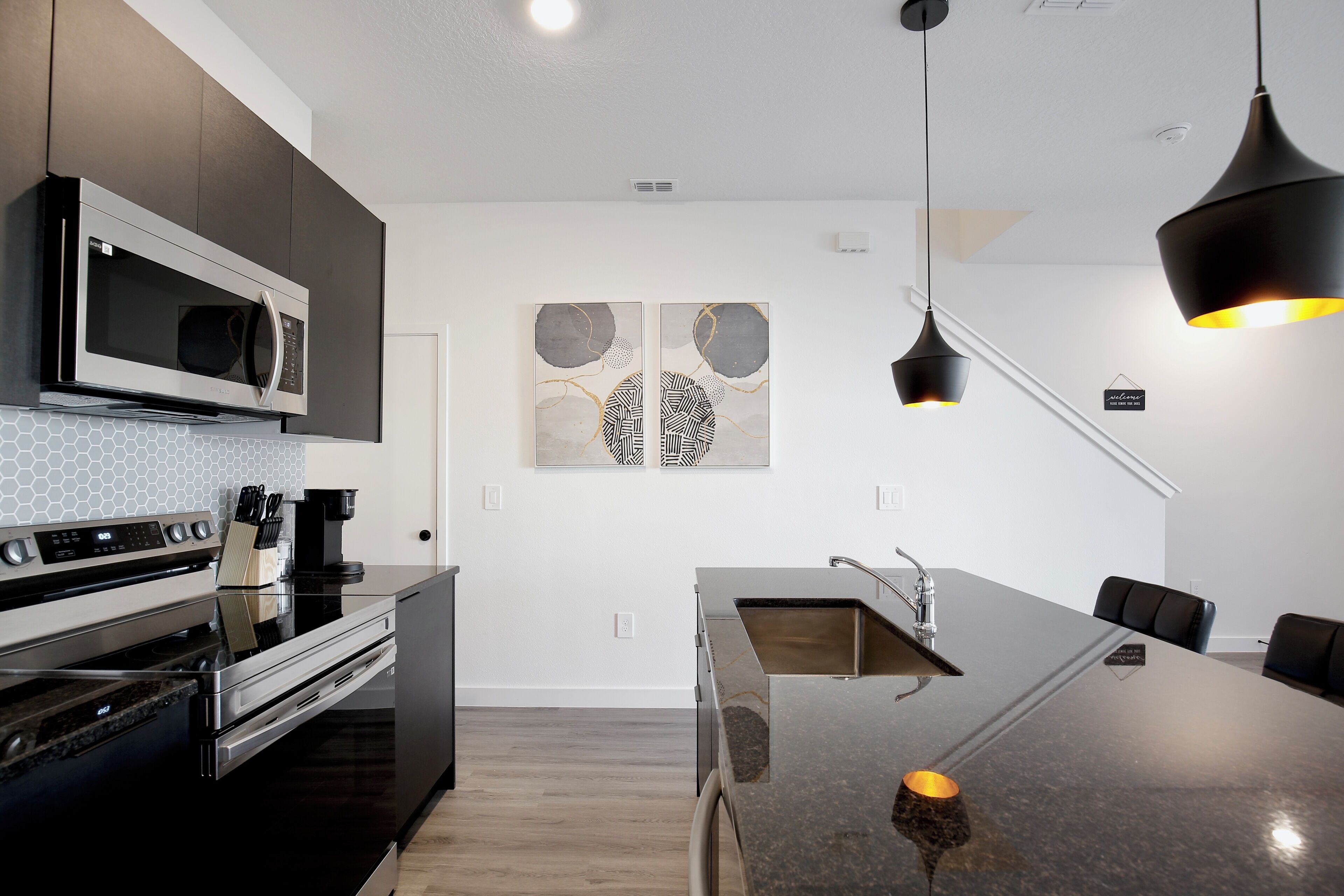 A modern kitchen with a built-in microwave, oven, and stove. There is a large island with a sink and a coffee machine. The kitchen has dark cabinets and a white backsplash.
