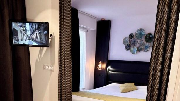 A hotel room with a bed, a television mounted on the wall, a bottle of wine, and a dining setup with plates, cutlery, and a plant. - Seclin Appartements (Seclin)