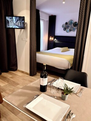 A hotel room with a bed, a television mounted on the wall, a bottle of wine, and a dining setup with plates, cutlery, and a plant. - Seclin Appartements (Seclin)