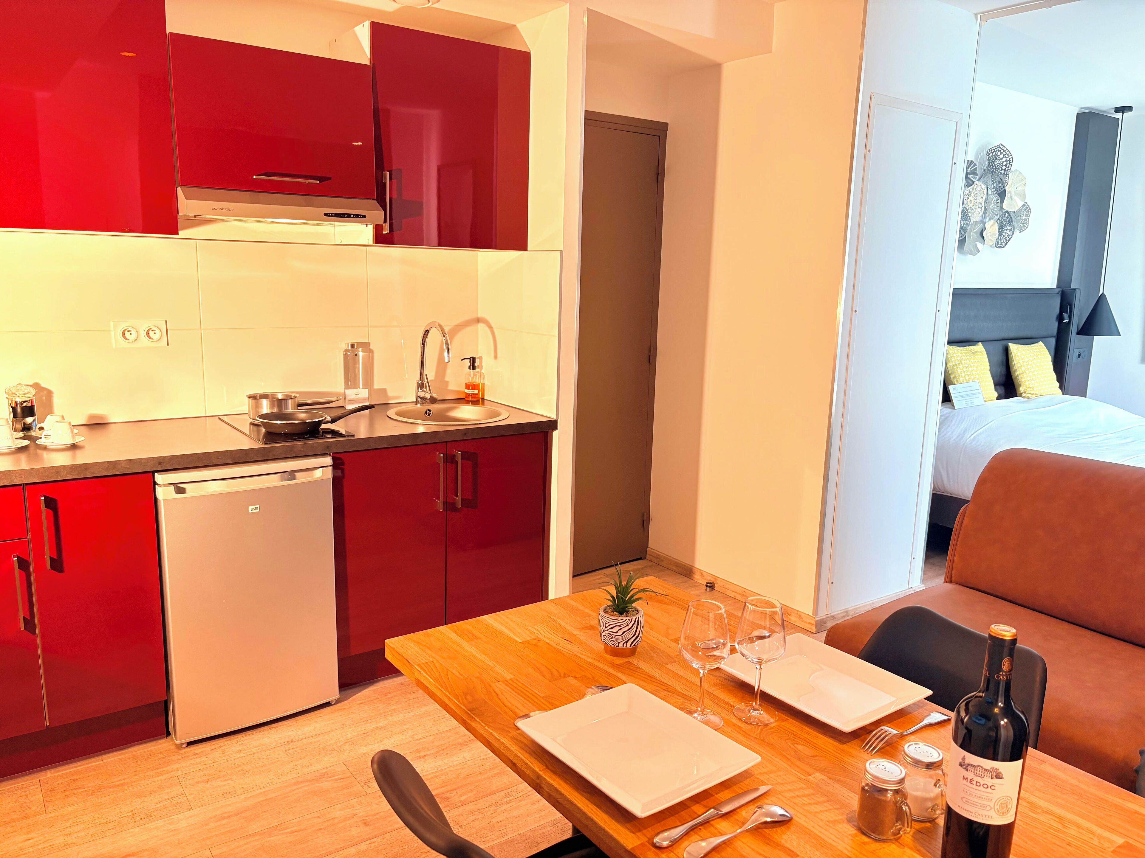 A compact kitchen with red cabinets, a dishwasher, and a dining area with a wooden table set for two.