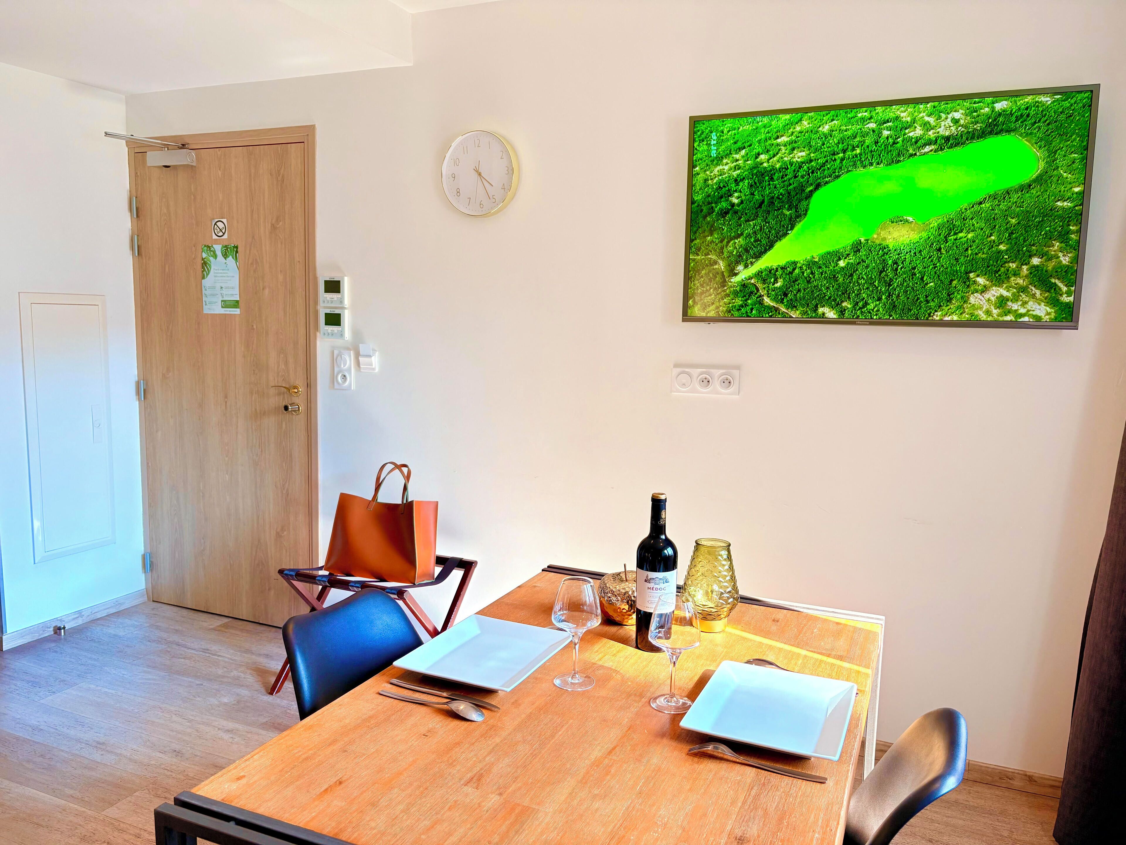 A dining area with a wooden table set for two, a laptop, wine glasses, and a bottle of wine. A clock and a television displaying a green landscape are on the wall.