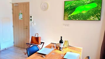 A dining area with a wooden table set for two, a laptop, wine glasses, and a bottle of wine. A clock and a television displaying a green landscape are on the wall.
