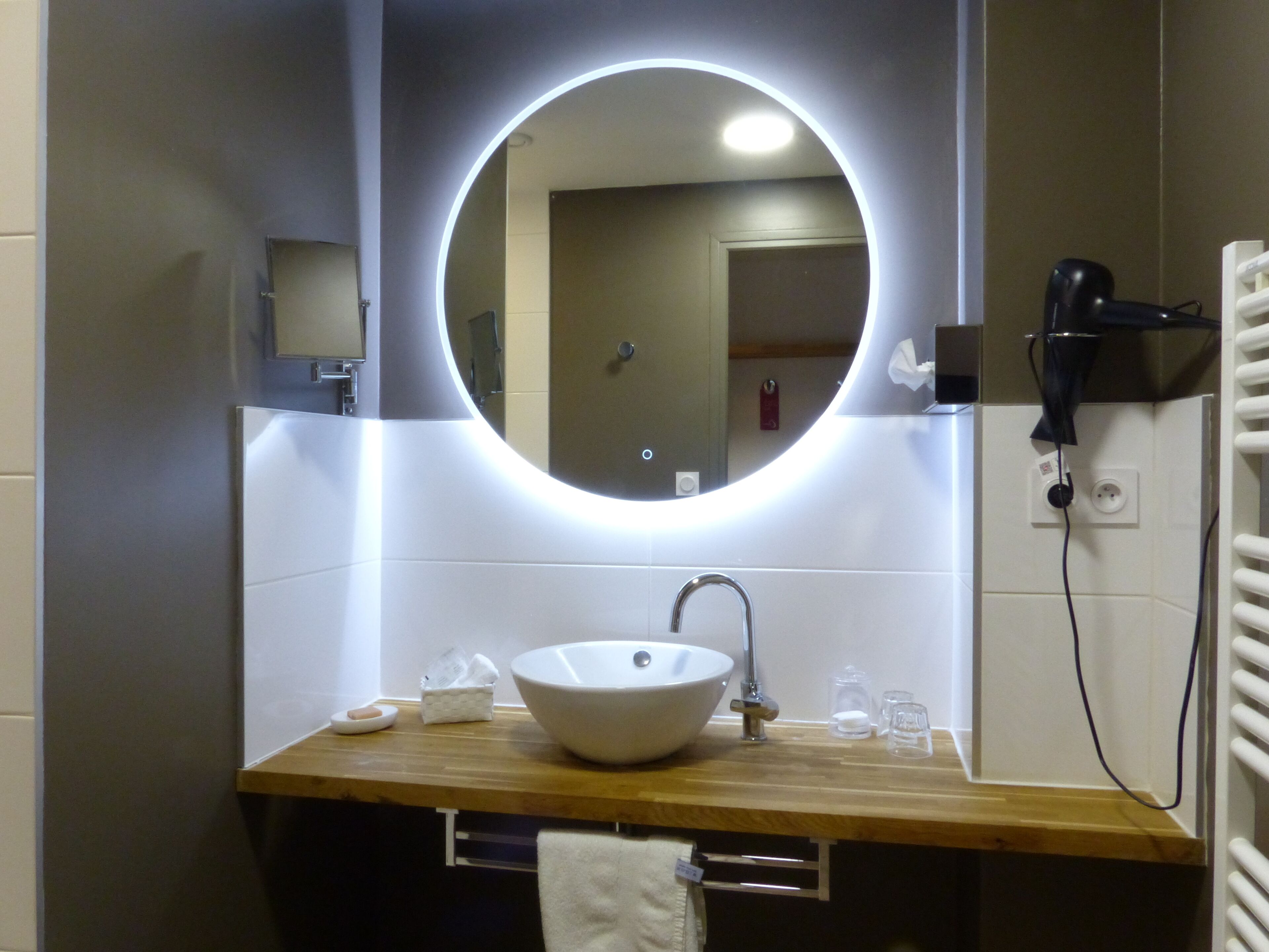 Un baño con un espejo redondo, un lavamanos blanco y un secador de pelo en un soporte de pared.
