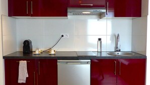 A compact kitchen with red cabinets, a dishwasher, a coffee maker, and a sink. - Seclin Appartements (Seclin)