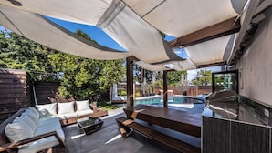 A modern outdoor patio with a pool, wooden furniture, and a covered seating area. - Luxury 3BR Oasis: Pool & Style (Costa Mesa)