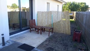 Een terras met een houten bankje en tafel, een glazen deur die naar een kamer leidt, en een bamboe hek.