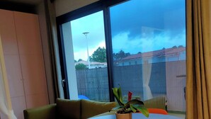 Een kamer met een ronde houten tafel, groene en oranje stoelen, een groene bank en een groot raam met uitzicht op een achtertuin.