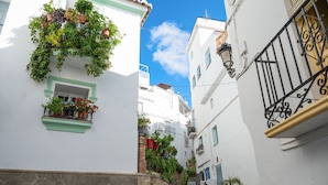 Une rue étroite bordée de bâtiments blancs, aux fenêtres ornées de cadres verts et aux balcons décorés de plantes en pot.