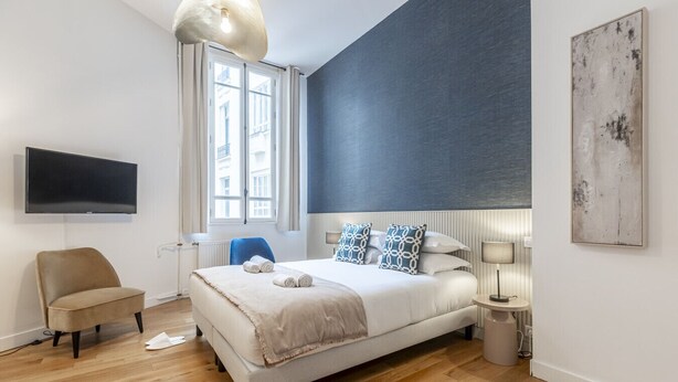 A modern bedroom with a large bed, a flat-screen TV, a chair, and a window with curtains. - Parc Monceau - Studio - 2 personnes (Paris)