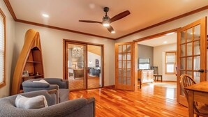A spacious living area with a wooden floor, a ceiling fan, and a view into the kitchen through glass doors.