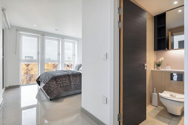 Une chambre avec un grand lit, une salle de bain avec des toilettes et un lavabo, et une vue sur le paysage urbain grâce à de grandes fenêtres.