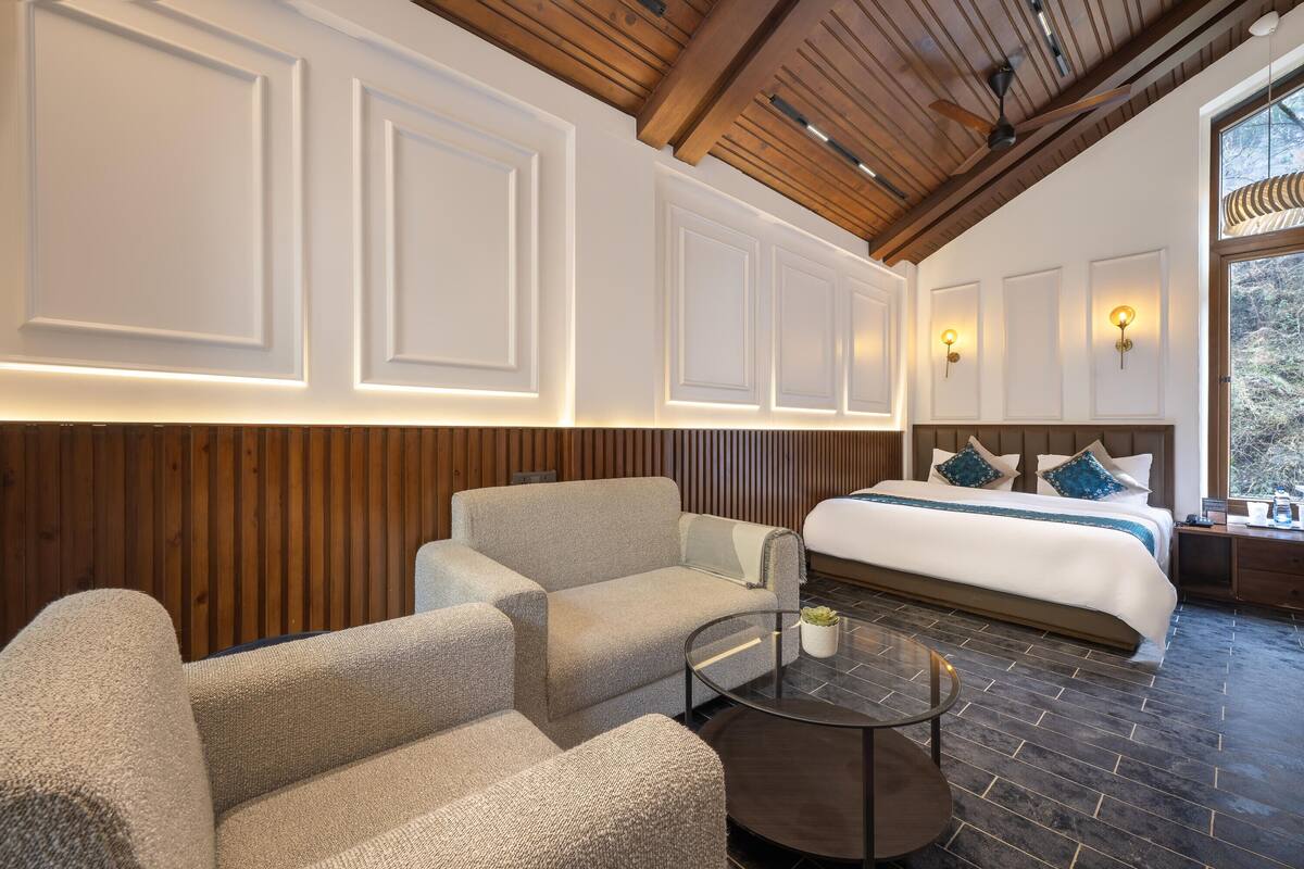 A modern hotel room with a large bed, two sofas, a round coffee table, and a view of the outdoors.