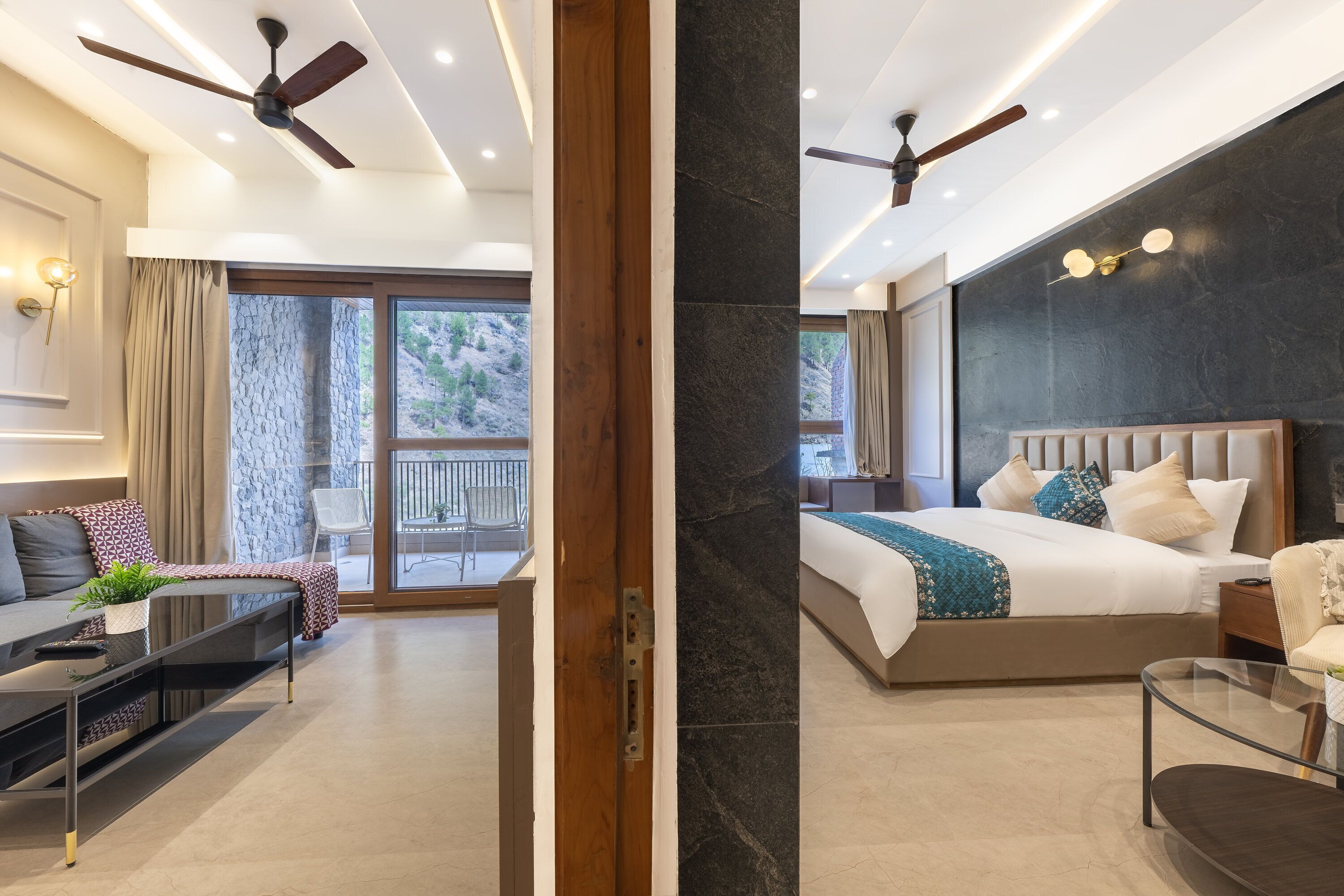 A modern hotel room with a bed, a sofa, a coffee table, and a balcony with a view of the mountains.