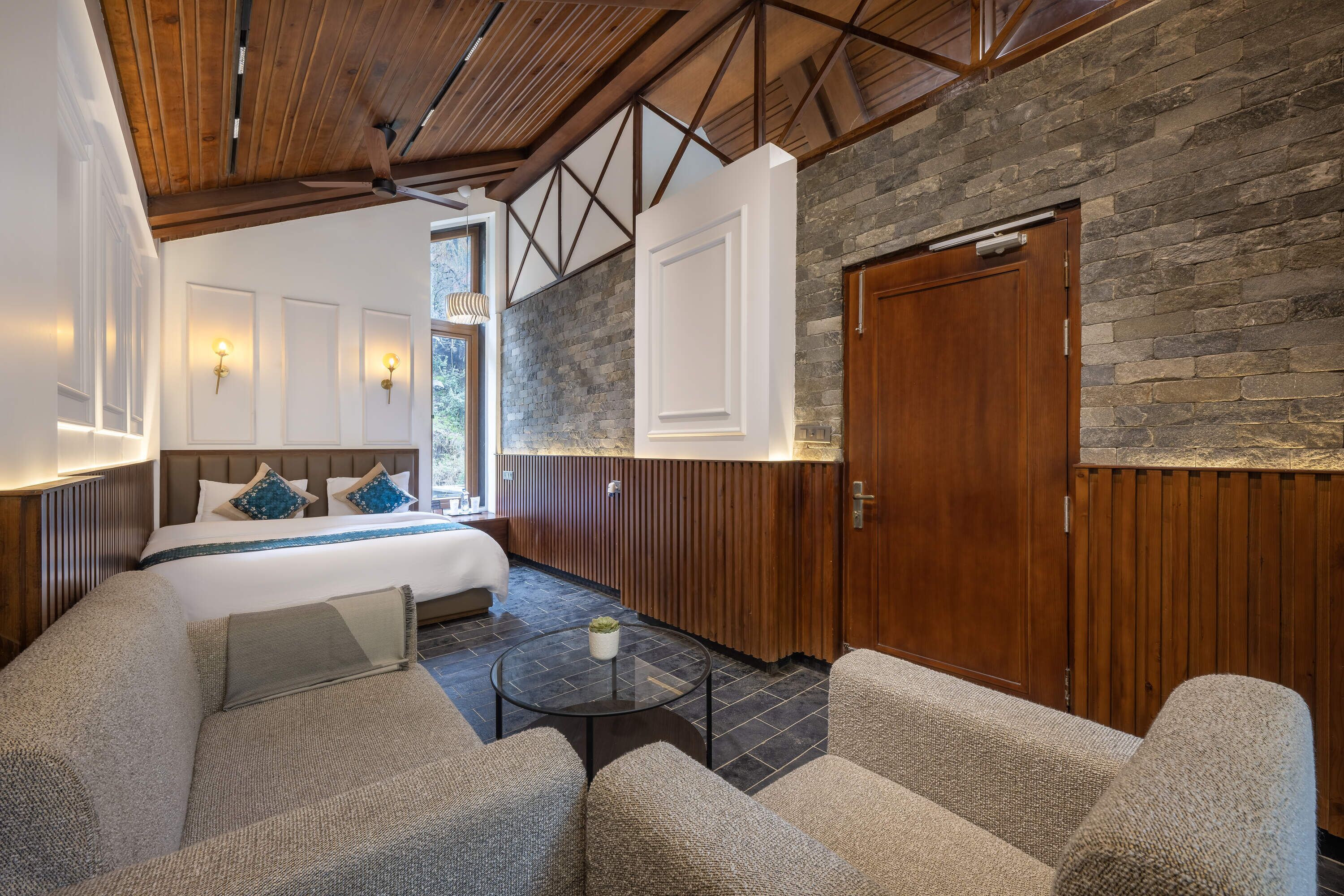 Una habitación de hotel moderna con techo de madera, paredes de piedra, una cama grande, un área de descanso con sofás y una pequeña mesa.