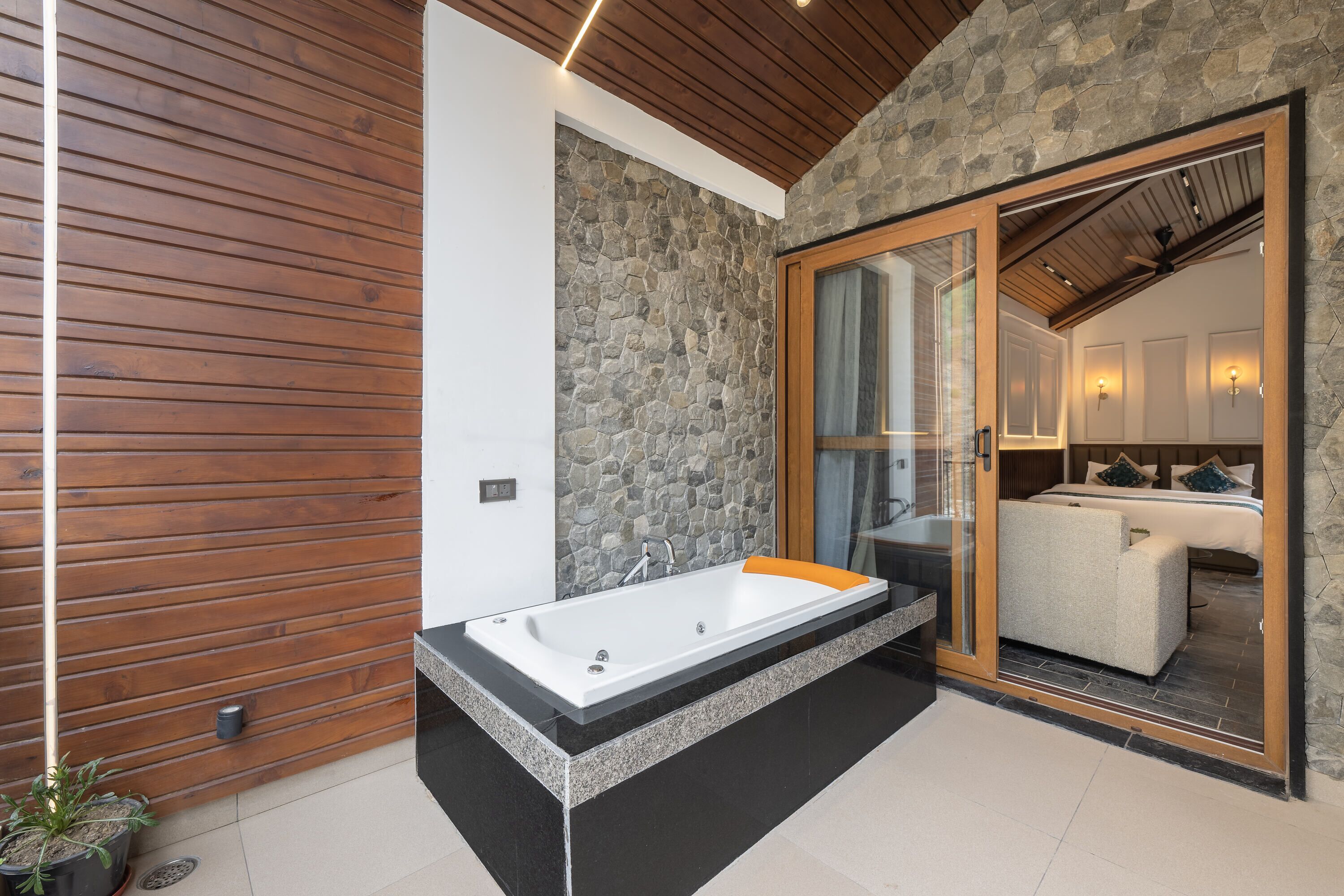 A modern bathroom with a stone wall, wooden paneling, a large bathtub, and a glass door leading to a bedroom.