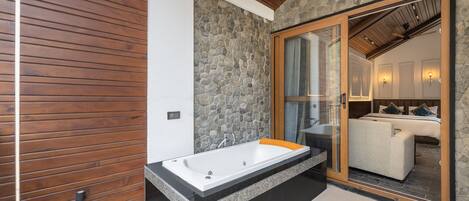 A modern bathroom with a stone wall, wooden paneling, a large bathtub, and a glass door leading to a bedroom.