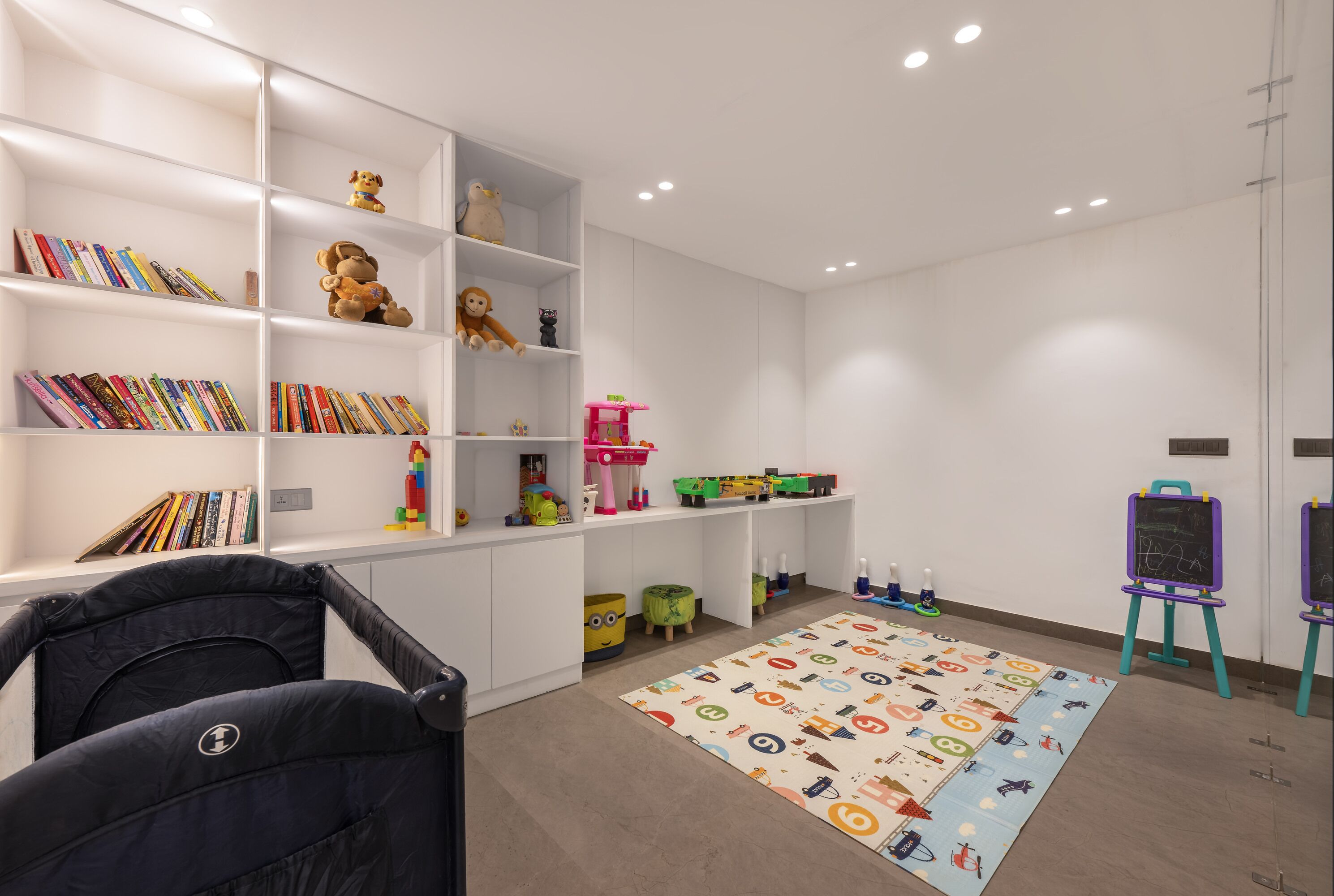 A room with white shelving units filled with books and toys, a colorful play mat, and a black chair.