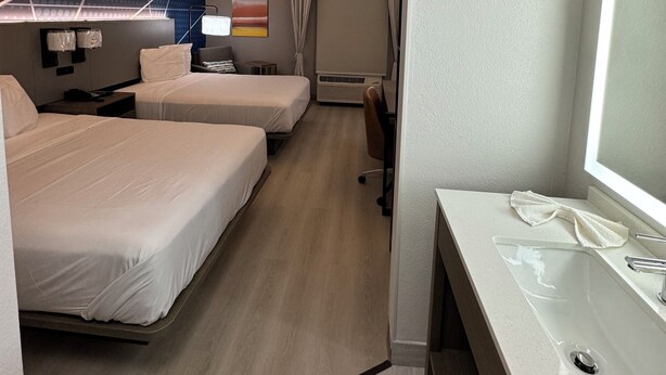 Une chambre d’hôtel avec deux lits, un bureau avec un miroir, une lampe de bureau, un téléviseur fixé au mur et une fenêtre avec des rideaux.