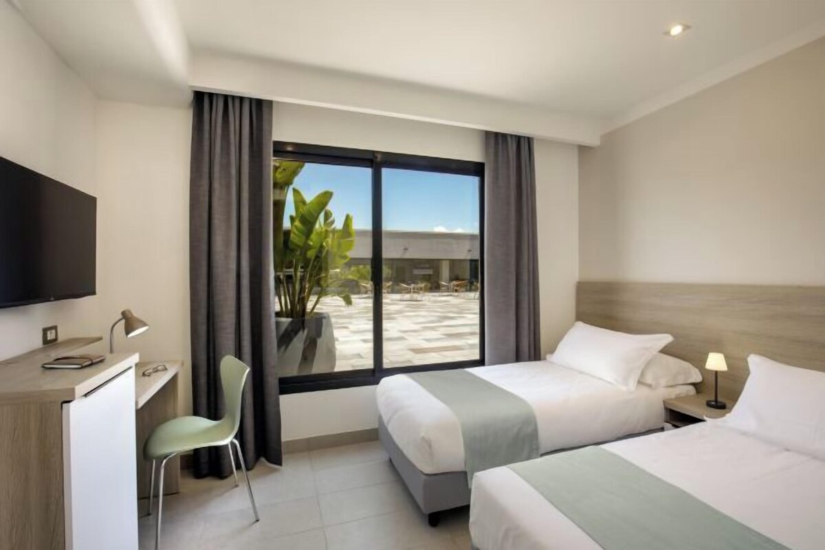 A hotel room with a desk, two beds, a TV, and a large window with curtains.