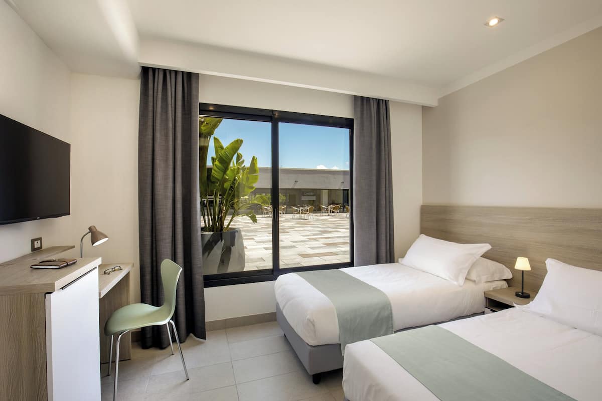 Habitación de hotel con dos camas, un escritorio, una silla, un televisor y un ventanal grande con cortinas.
