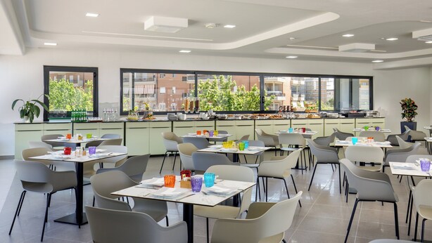 A modern dining area with multiple tables and chairs, large windows with a view of buildings and trees, and a clean, contemporary design. - SIMON HOTEL APRILIA (Aprilia)
