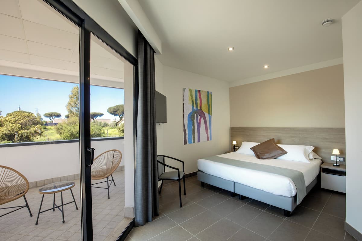 Habitación de hotel moderna con una cama grande, un balcón con sillones de mimbre y vistas al exterior.