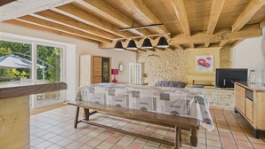 A bedroom with a wooden ceiling, a bed with a patterned bedspread, a stone wall, a television, and a window with a view of greenery.