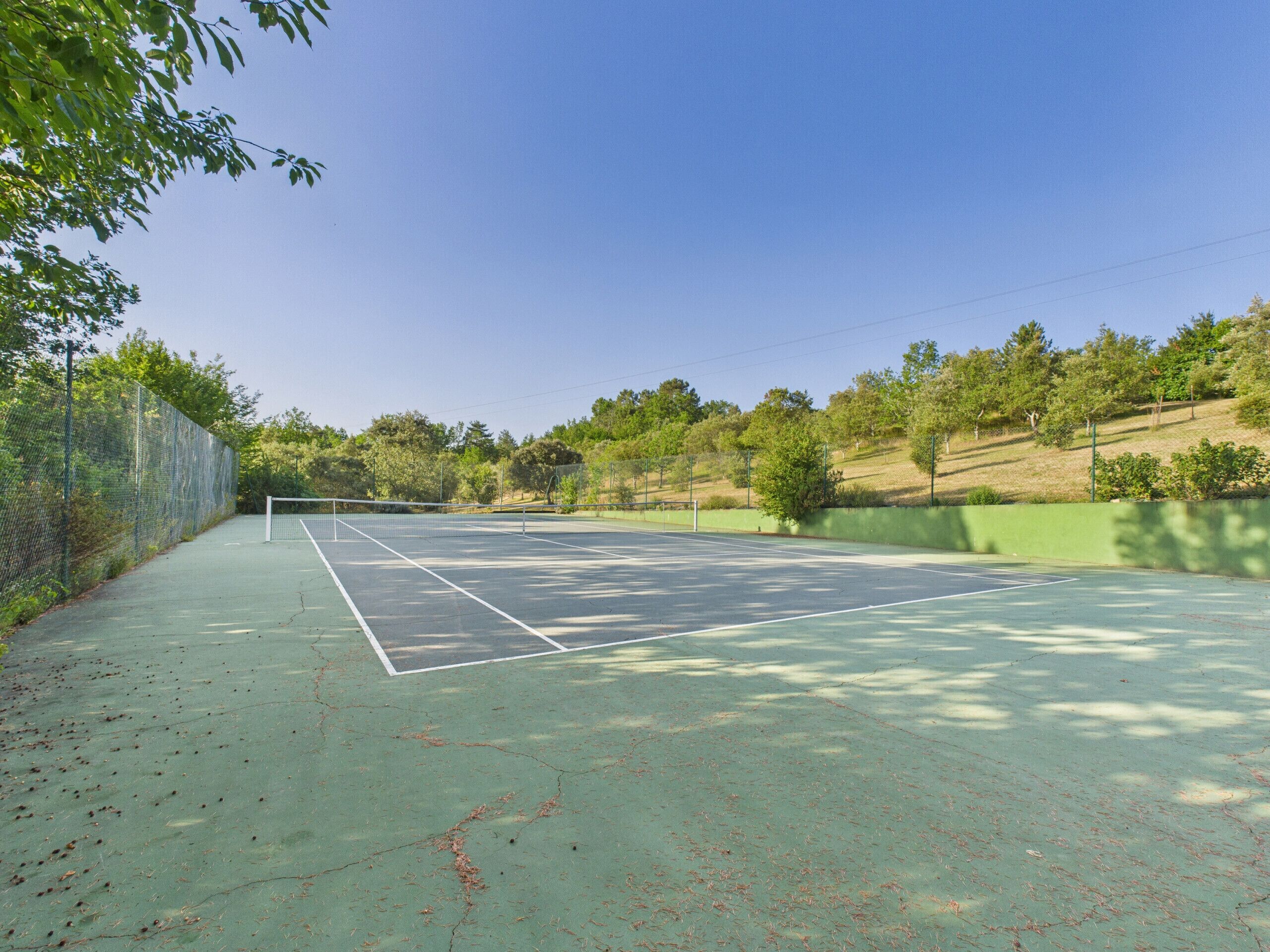 An outdoor tennis court with a net, surrounded by trees and a green fence.