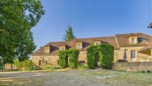 A stone building with a gravel driveway, a covered patio, and a large tree to the left.