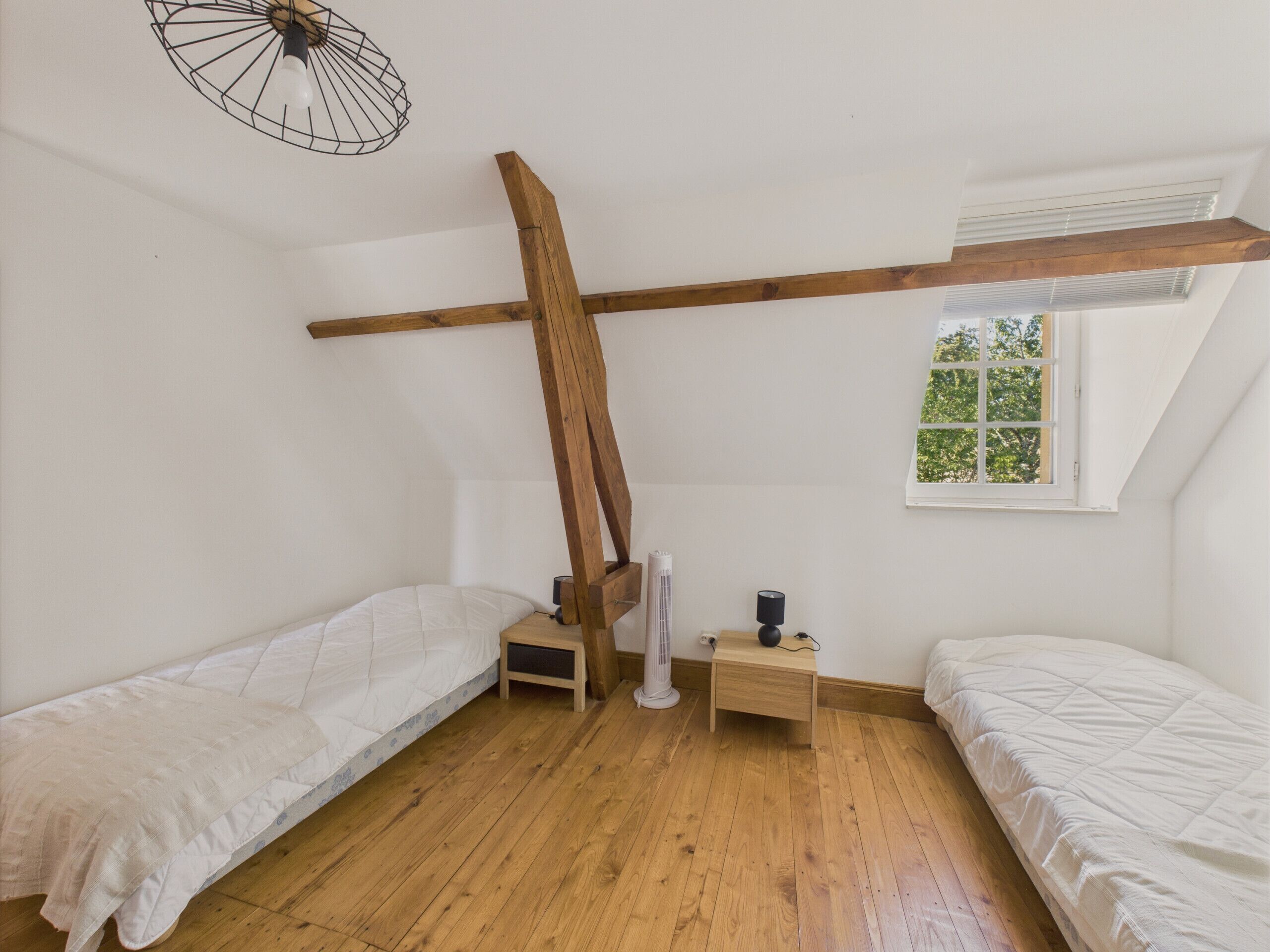 A room with two single beds, wooden beams, a small table, a fan, and a window with a view of greenery.