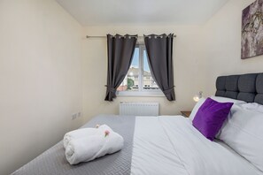 A bedroom with a bed, a window with curtains, a radiator, a nightstand with a lamp, and a wall-mounted artwork.