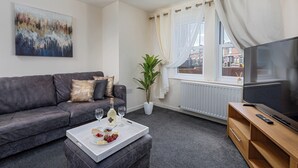 Ein modernes Wohnzimmer mit einem grauen Sofa, einem Couchtisch mit Snacks, einer Topfpflanze, einem Fernseher und einem Fenster mit Vorhängen.