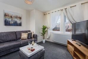 A modern living room with a grey sofa, a coffee table with snacks, a potted plant, a TV, and a window with curtains. - House - Sleeps 10 - Parking - Wifi (Barrow-in-Furness)