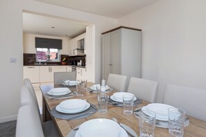 A dining area with a wooden table set for four, white chairs, and a kitchen in the background. - House - Sleeps 10 - Parking - Wifi (Barrow-in-Furness)