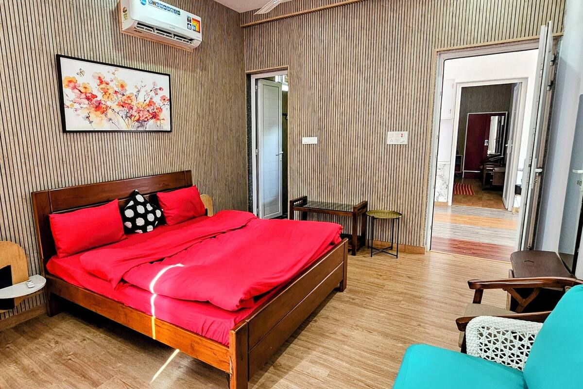 A hotel room with a wooden bed, red bedding, a blue sofa, a small table, and a wall-mounted air conditioner.
