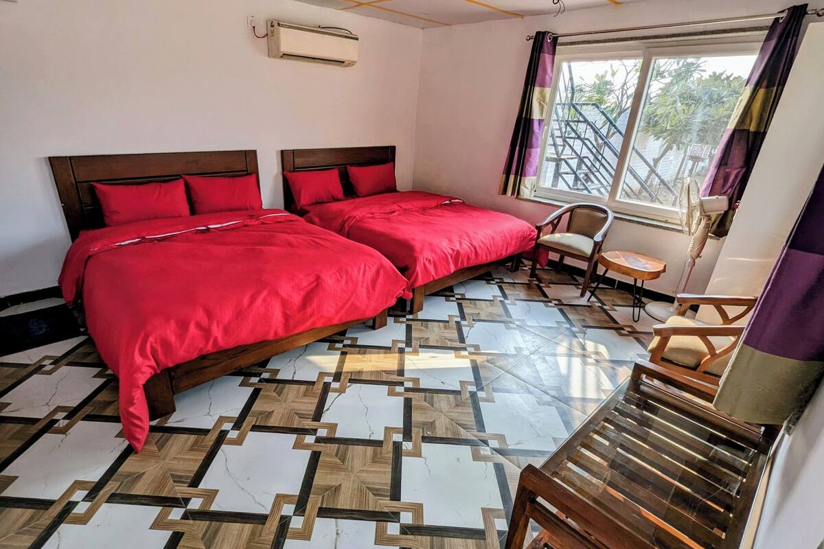 A room with two beds, a small table, a chair, and a window with a view of greenery.