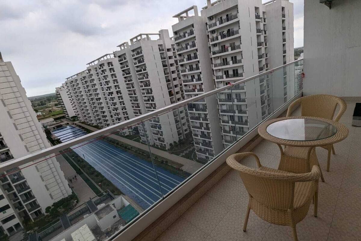 A balcony with wicker furniture and a glass table, overlooking a swimming pool and high-rise buildings.