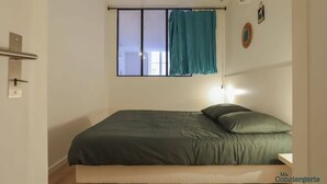 A single bed with a green mattress and pillows, a bedside table, a window with a curtain, and a wall-mounted light fixture.