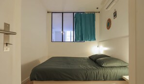 A single bed with a green mattress and pillows, a bedside table, a window with a curtain, and a wall-mounted light fixture.