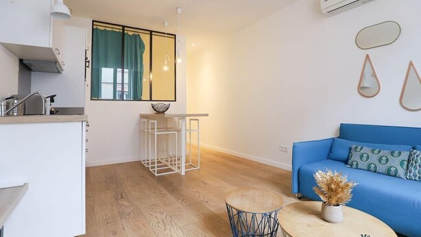 A modern living room with a blue sofa, a small round table with a vase, and a kitchen area with a window and a mirror.