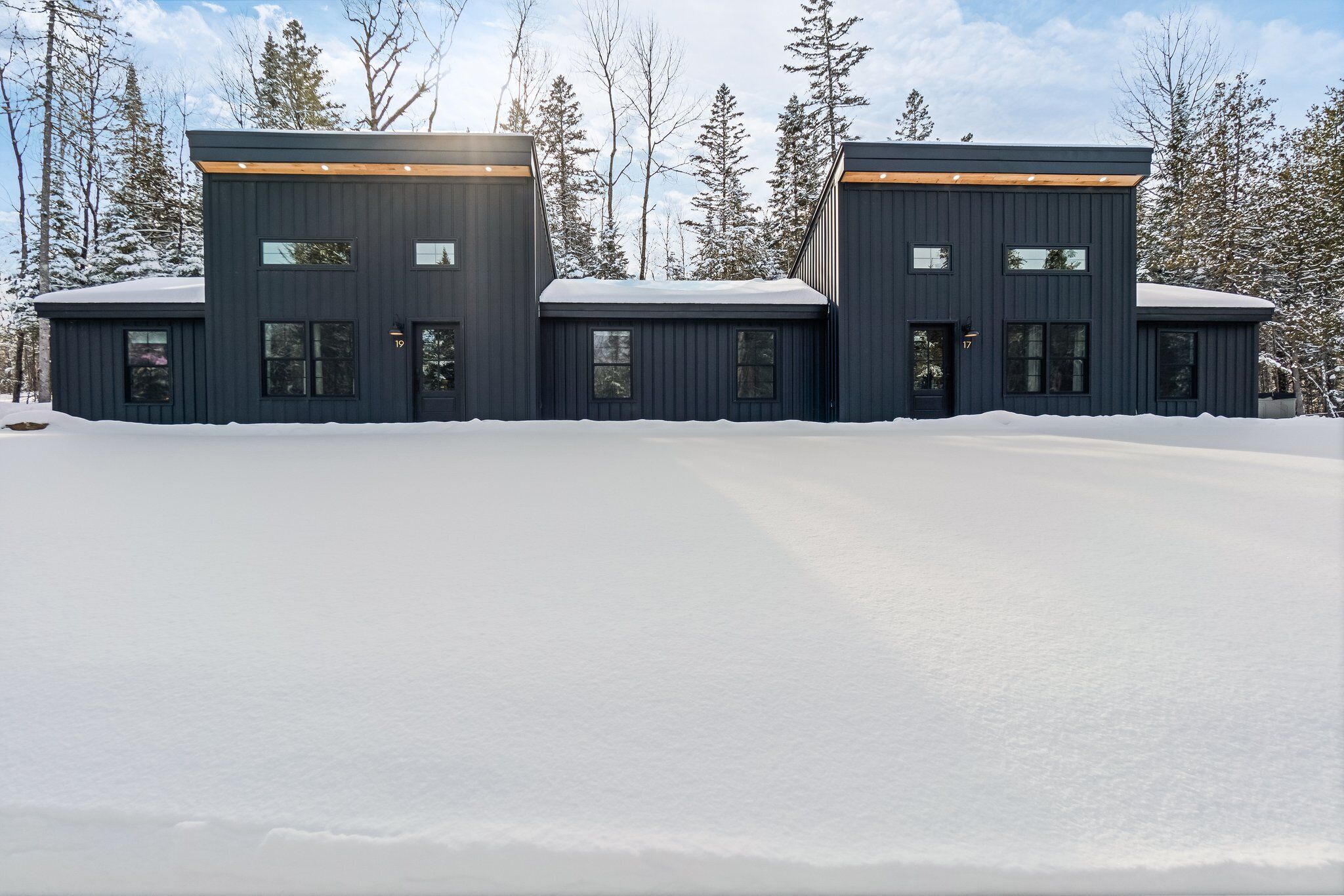 Un bâtiment moderne de couleur sombre, doté de nombreuses fenêtres et d’un toit plat, entouré de neige et d’arbres.