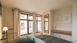 Une chambre d’hôtel comprenant un lit, une table de chevet avec une lampe, une fenêtre avec des rideaux et un mur orné de deux tableaux encadrés.