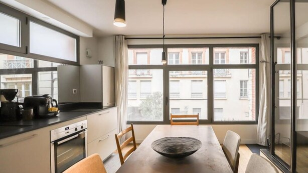 Une cuisine moderne avec une grande fenêtre, une table à manger agrémentée d’un centre de table décoratif, et une vue sur les bâtiments environnants.