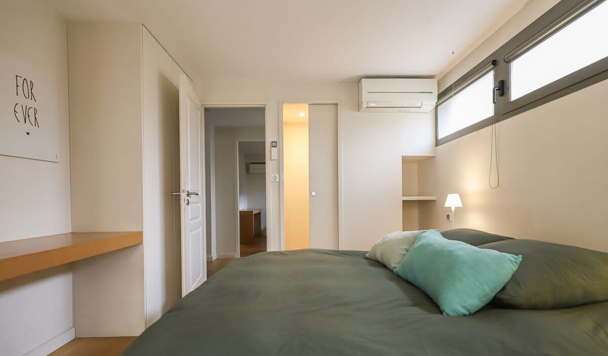 A bedroom with a bed, a bedside lamp, a window, and a door leading to another room.