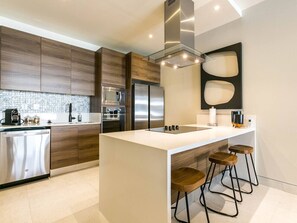 A modern kitchen with a central island, bar stools, stainless steel appliances, and a backsplash. - Brand-new, modern, 2-bedroom, near Cabo Marina (Cabo San Lucas)