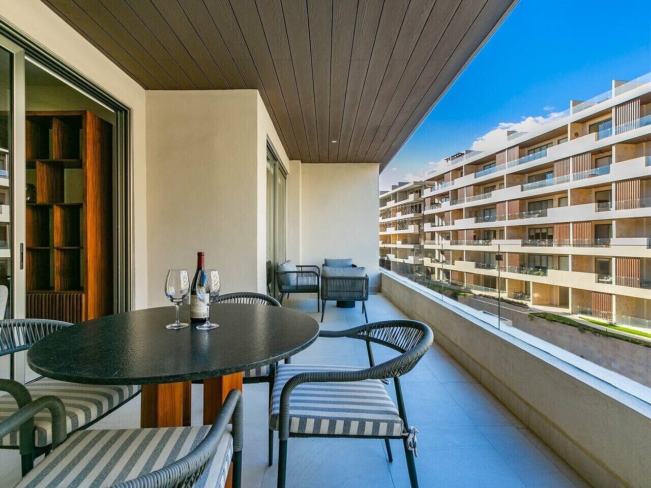 A balcony with a round table and chairs, a wine bottle, and a view of a multi-story building.