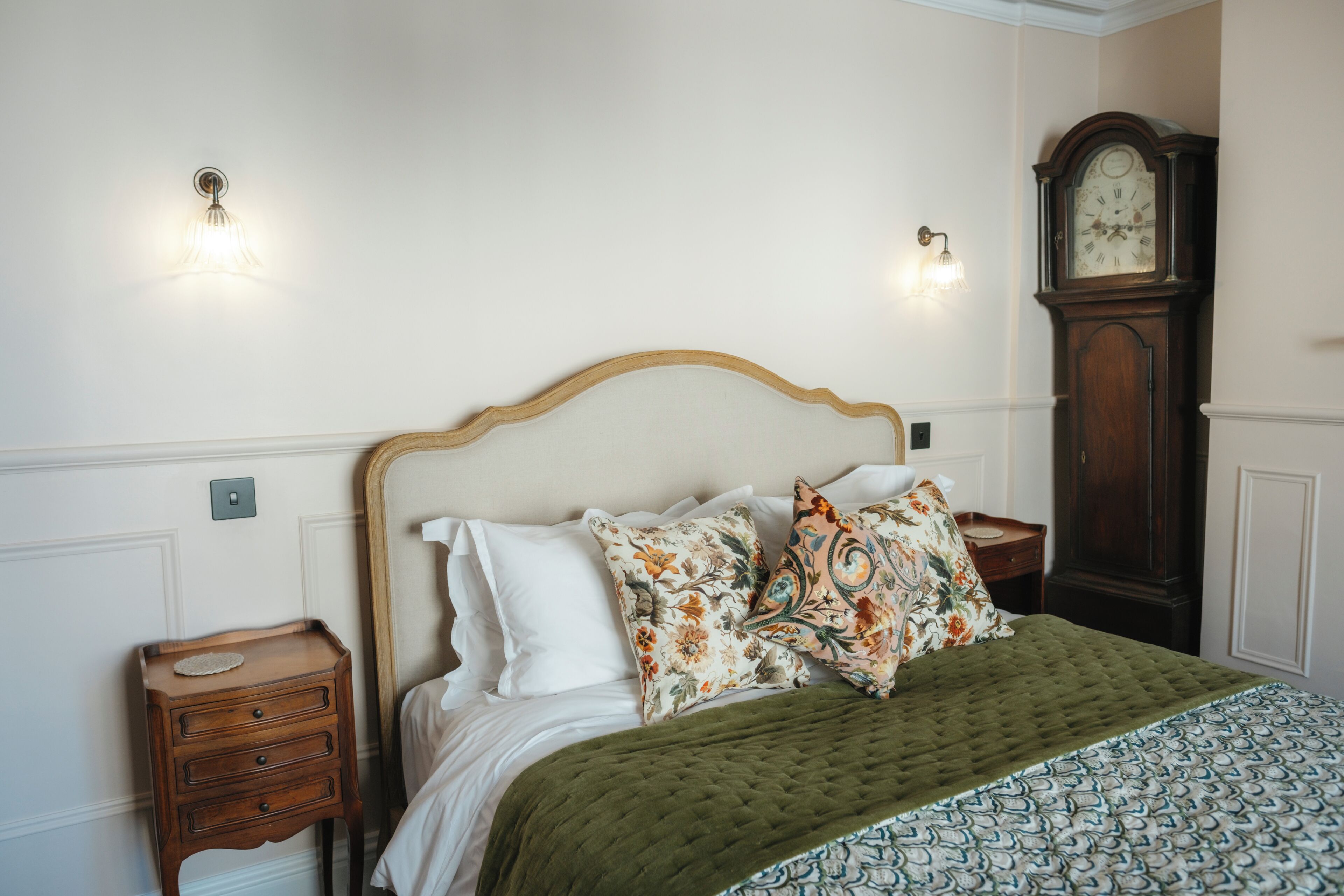 Una cama bien hecha con un edredón verde, dos almohadas florales y una mesita de noche de madera con cajones. Se ve un reloj de pared a la derecha.