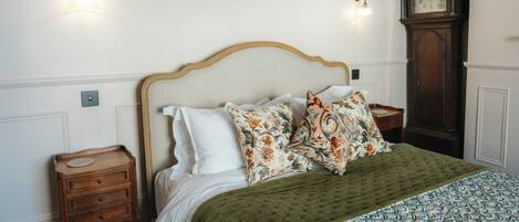 Una cama bien hecha con un edredón verde, dos almohadas florales y una mesita de noche de madera con cajones. Se ve un reloj de pared a la derecha.