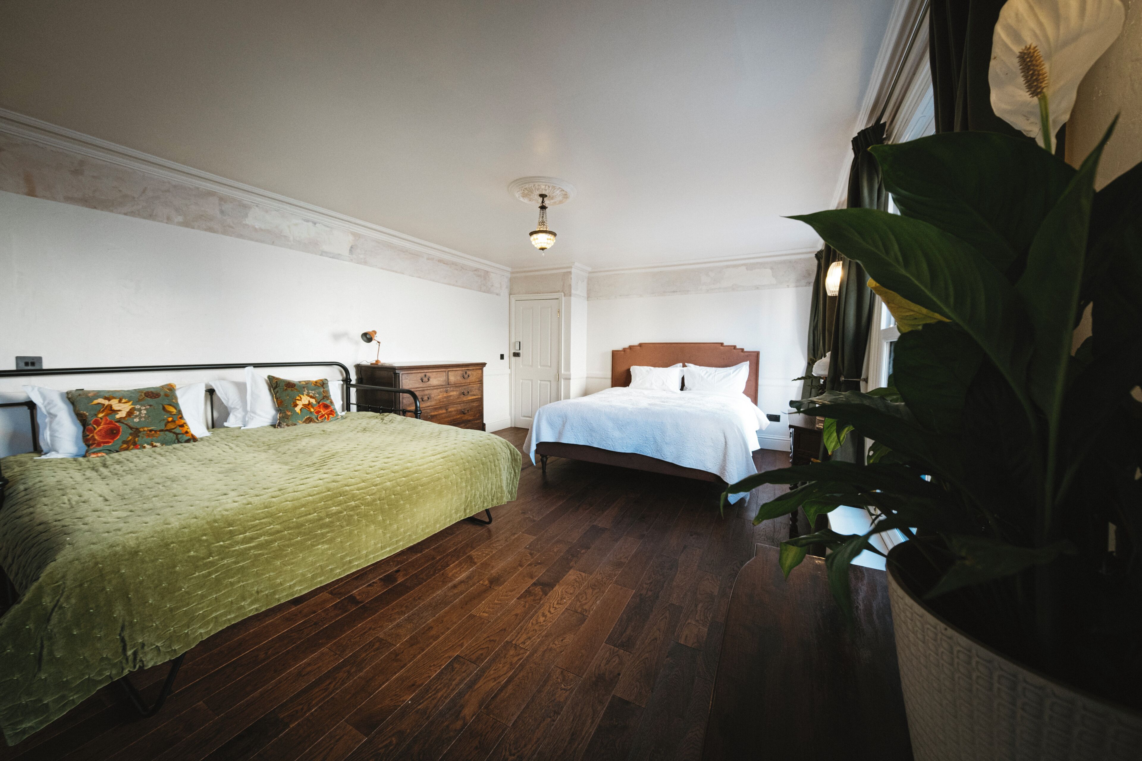 A bedroom with two beds, wooden flooring, a large potted plant, and a classic ceiling light fixture.