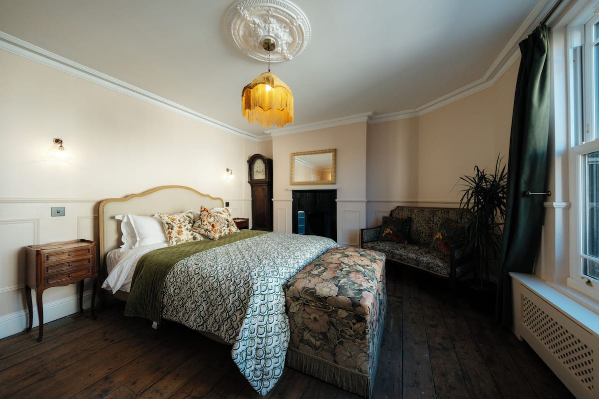 A bedroom with a bed, a nightstand, a bench, a mirror, a clock, and a window with curtains.